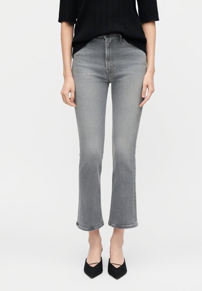 Γυναίκα που φοράει γκρι τζιν ψηλόμεσο cropped με καμπάνα, μαύρο τοπ και μαύρες μυτερές γόβες, στέκεται μπροστά σε απλό φόντο.