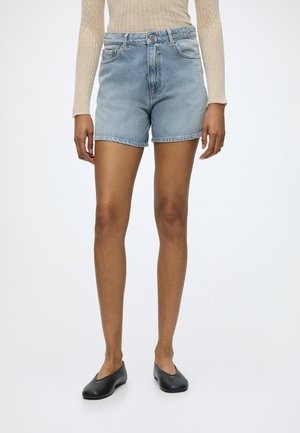 Person trägt hellblaue Jeansshorts, beige langärmelige gerippte Oberteile und schwarze flache Schuhe, steht vor einem schlichten weißen Hintergrund.