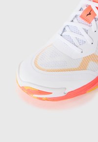 White athletic shoe front with mesh upper, white laces, and orange-pink gradient sole detail on a light background.