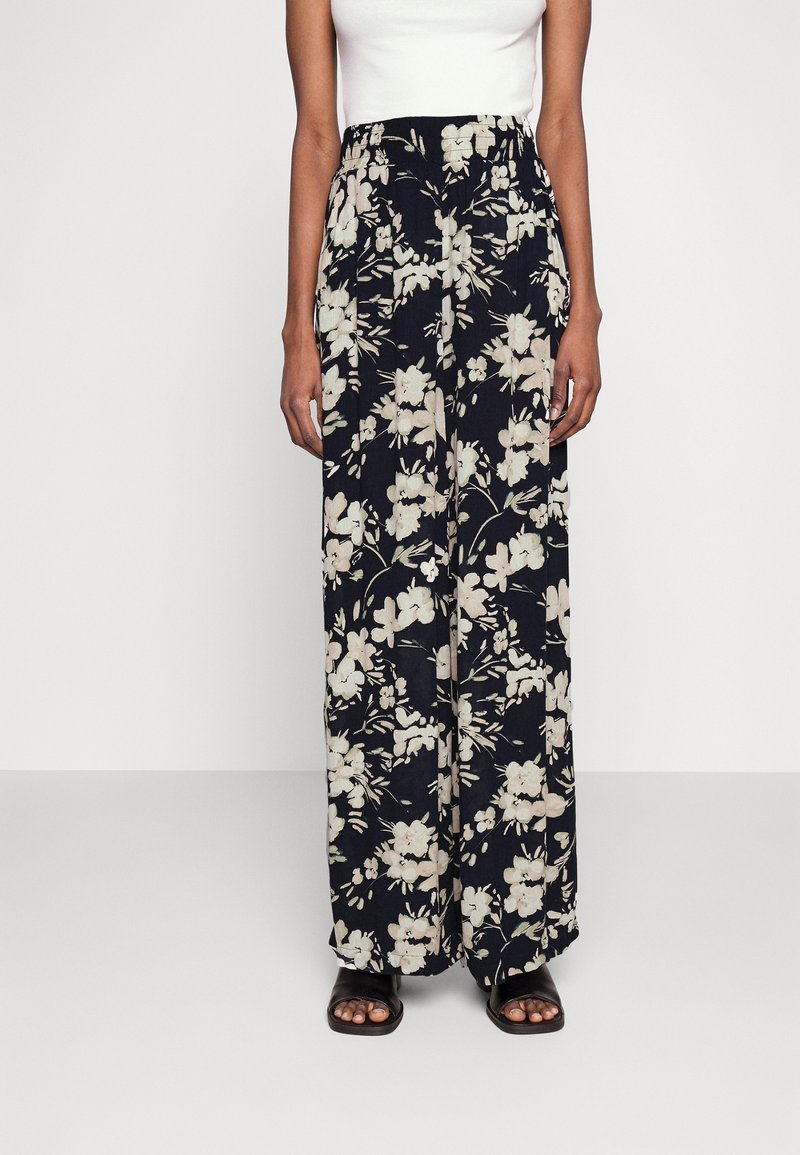 Pantalon large à motif floral marine, avec un motif floral léger. Taille élastique et tissu fluide pour plus de confort. Porté avec un haut blanc et des sandales.