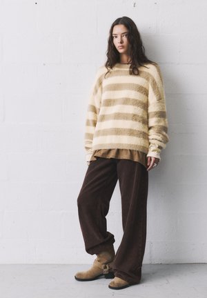 Gestreifter, fester Pullover in Creme und Tan, über einem braunen Ripp-Shirt getragen, kombiniert mit weiten, braunen Cordhosen und beigen Ankle Boots.
