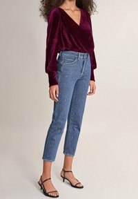 Bordeaux fluwelen wikkeltop met lange mouwen, gecombineerd met hooggeknoopte blauwe jeans met een stippenversiering. Zwarte gespsandalen.