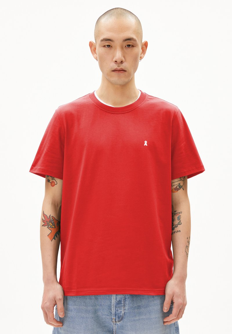 T-shirt rouge à manches courtes en coton avec un col rond. Présente un petit détail en ruban blanc sur la poitrine gauche. Porté avec un short en denim bleu.