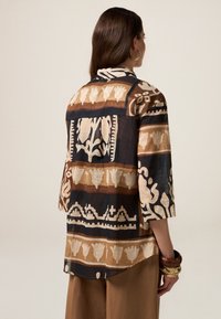Langarmshirt in Schwarz, Creme und Braun mit gemustertem Design, das botanische Motive zeigt und einen offenen Rücken hat. Leichter, strukturierter Stoff.