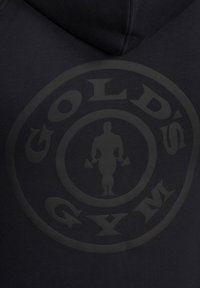 Ciemna bluza z kapturem z okrągłym logo Gold’s Gym, na którym w centralnej części znajduje się muskularna postać trzymająca hantle.