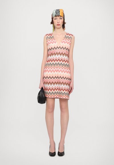 Knit dress with a zigzag pattern in pink, red, and brown. Features a deep V-neck, sleeveless design, and a fitted silhouette. Black accessories.