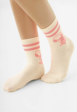 Voeten met lichtbeige sokken met roze strepen en Snoopy-figuurtjes op een effen witte achtergrond.