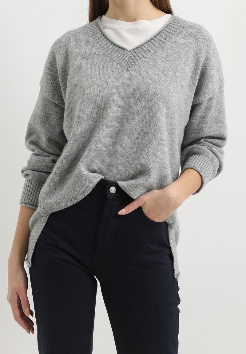 Suéter gris de cuello en V hecho de una suave tela de punto. Presenta puños y dobladillo de canalé, aberturas laterales, usado sobre una camisa blanca. Combinado con pantalones oscuros.