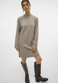 Beiges Strickkleid mit langen Ärmeln und hohem Kragen, mit gerippten Akzenten. Kombiniert mit hohen, braunen Lederstiefeln. Schlichtes Design.