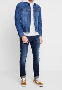 Man med blå denimjacka, ljusgrå hoodie, mörkblå jeans och bruna mockaskor med vita snören, stående mot en vit bakgrund.