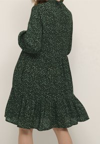 Vestido floral verde con mangas largas, cintura fruncida y bajo con volantes. Material ligero con un pequeño patrón de hojas en varios tonos de verde.