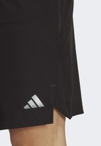 Pantalones cortos deportivos negros hechos de una tela suave con un logo de Adidas reflectante en blanco cerca del dobladillo y un bolsillo con cremallera en el lateral.