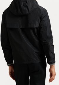 Musta hupullinen takki, jossa on matta pinta ja kevyt kiilto. Takissa on takalahje ja resoriset hihansuut, suunniteltu väljäksi istuvuudeksi.