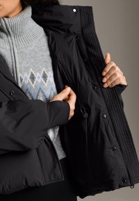 Čierna zateplená bunda s patentkami, vreckami na zips a rebrovanými manžetami, navrchu svetlosivého svetra s modrými a sivými diamantovými vzormi.