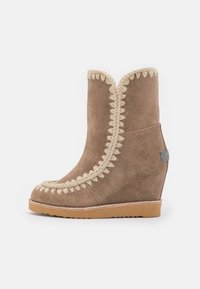 Botín cuña de ante marrón con costuras decorativas en crema a lo largo del borde, puntera redondeada y un pequeño detalle de logo en el tacón.