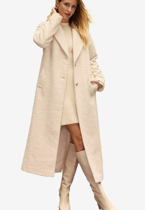 Long manteau en laine crème avec un col châle, deux poches avant et un tissu texturé, associé à une robe crème à côtes et des bottes jusqu'aux genoux.