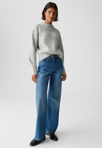 Frau steht in einem hellgrauen gerippten Pullover, hochtaillierten, weiten blauen Jeans mit einem schwarzen Gürtel und schwarzen Schuhen vor einem schlichten Hintergrund.
