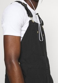 Salopette nera con tasca frontale, dettagli in metallo dorato e bretelle regolabili, indossata su una t-shirt bianca. Tessuto liscio, design informale.