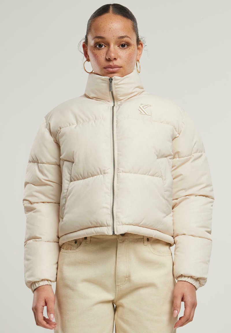 Lys beige cropped puffer-jakke med høy krage, glidelås foran og elastiske mansjetter. Har en subtil logo på venstre bryst.