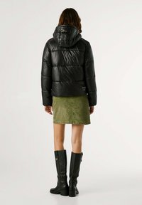 Zwarte pufferjas met een capuchon, met een gequilt ontwerp. Gepaard met een groene suède rok en kniehoge zwarte haklaarzen.