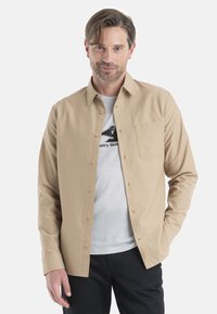Camicia beige con bottoni realizzata in un tessuto leggero, con taschino sul petto, maniche lunghe e colletto classico, indossata sopra una maglietta chiara.
