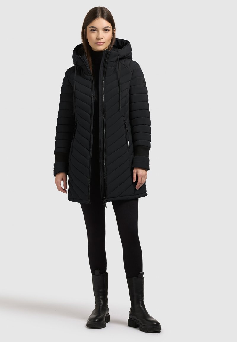 Black puffer coat with a chevron pattern, hood, zipper closure, and side pockets. Worn over black leggings and ankle boots.