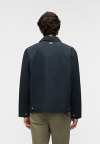 Μπουφάν σε ναυτικό μπλε χρώμα με γιακά, μακριά μανίκια και μικρό κεντημένο λογότυπο στο πίσω μέρος. Διαθέτει λεπτομέρειες με πιέτες στο τελείωμα.