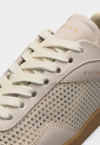 Baskets beiges avec panneaux latéraux en mesh texturé, lacets crème, détails surpiqués et logo "NUBIKK" sur la languette et le côté.