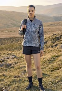 Noor isik, kes kannab halli mustrilist jopet, musti lühikesi pükse ja matkasaapaid, seisab rohelisel künkal, seljas on seljakoti rihm.