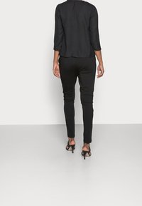 Schwarzes langärmeliges Oberteil kombiniert mit figurbetonten schwarzen Hosen und schwarzen High-Heel-Sandalen, von hinten vor einem neutralen Hintergrund gesehen.