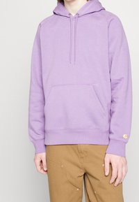 Person som bär en lavendeltröja med framficka och beige byxor, stående mot en enfärgad bakgrund.