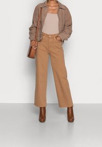 Giacca scamosciata tan, canotta beige a costine, pantaloni larghi marrone chiaro e stivali con tacco marroni. Una borsa a spalla marrone completa l'outfit.