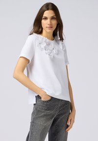 Witte korte mouwen shirt met een getextureerde volantsachtige halslijn en zilveren accenten, gecombineerd met grijze jeans met studdetails.