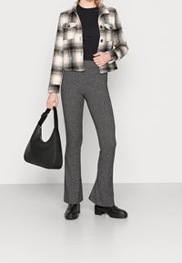 Karierte Flanelljacke in Schwarz und Weiß, enganliegendes schwarzes Oberteil, graue strukturierte Schlaghose, schwarze Umhängetasche und klobige schwarze Stiefel.