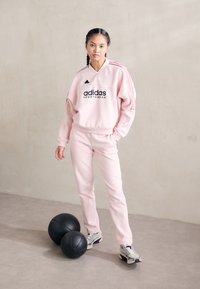 Roze Adidas sportoutfit met zwart logo en drie witte strepen, gecombineerd met grijze en paarse sportschoenen, naast twee zwarte ballen.