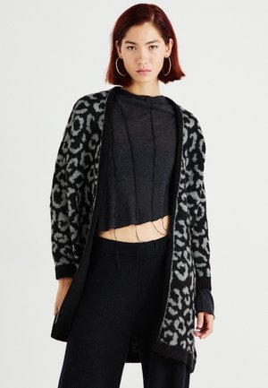 Cardigan à imprimé léopard noir et gris en matériau tricot doux, avec des manches longues et un design ouvert, superposé à un top noir court.