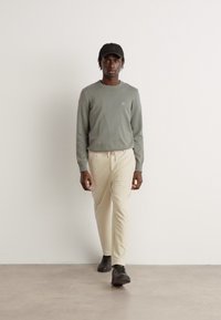 Chemise à manches longues gris clair avec un petit logo en patch, pantalon crème et baskets noires. Modèle portant une casquette noire, se tenant sur un sol neutre.