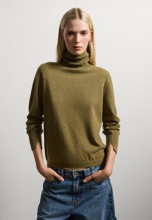Maglione a collo alto verde oliva in morbido materiale a maglia, con maniche lunghe dotate di polsini a coste e fessure laterali, abbinato a pantaloni shorts in denim a vita alta.
