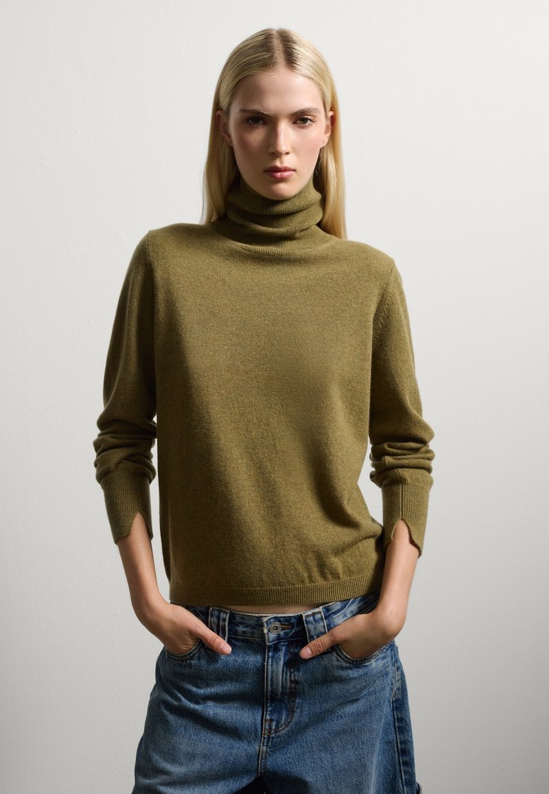 Olivgrüner Rollkragenpullover aus weichem Strickmaterial, mit langen Ärmeln und gerippten Bündchen sowie Seitenschlitzen, kombiniert mit hochgeschnittenen Jeansshorts.