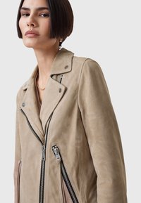 Beige suède bikerjack met dubbele rits, drukknoopkleppen en contrasterende zijkanten. Soepele textuur en gestructureerde pasvorm.