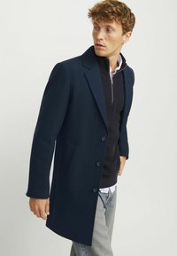 Abrigo azul marino con un diseño ajustado, hecho de una tela estructurada. Presenta un cuello con muescas, tres botones y bolsillos laterales. Llevado sobre un suéter azul marino.