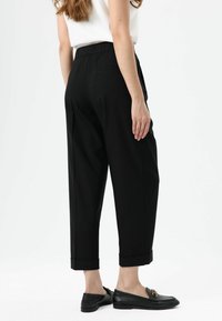 Zwarte getailleerde broek met een elastische tailleband, taps toelopende pijpen en opgerolde zoom, gecombineerd met zwarte loafers met gouden accenten.