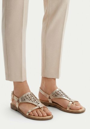 Voeten met beige sandalen met decoratieve uitsnijdingen en een kleine hak, gecombineerd met lichtbeige broeken die boven de enkel zijn afgeknipt.