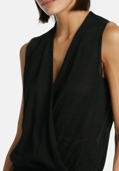 Femme portant un haut noir sans manches drapé avec un décolleté en V profond, montrée des épaules jusqu'au milieu du torse sur un fond uni.