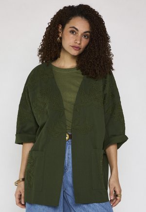 Femme aux cheveux bouclés portant une veste brodée de fleurs vert foncé, une chemise vert olive, un jean bleu et des accessoires dorés sur un fond clair.