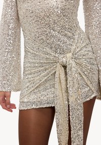 Robe à sequins en or métallique avec manches longues. Dotée d'un détail de taille nouée avec un tissu fluide, créant un aspect texturé et superposé.
