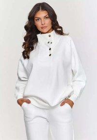 Biała bluz sweatshirt z wysokim kołnierzem i zapięciem na trzy guziki. Miękki materiał, luźny krój i żeberkowe mankiety. Noszona z dopasowanymi spodniami.