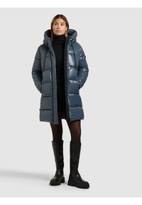 Kvinna som bär en lång, blå vadderad huva jacka, svart polotröja, kjol, strumpbyxor och knähöga svarta stövlar, stående med händerna i fickorna.