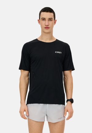 Man wearing black short-sleeve athletic shirt with "cep" logo and light gray running shorts, standing against white background.