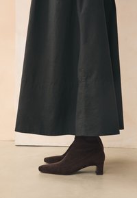 Stivaletti alla caviglia in camoscio marrone con punta appuntita e tacco basso a blocco, abbinati a una gonna midi in tessuto nero, creando un look lineare.